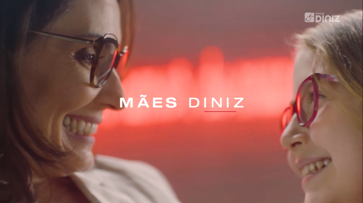 Mulher usando óculos olha, sorridentes para uma menina, que também está de óculos. Celebração do Mês da Mães